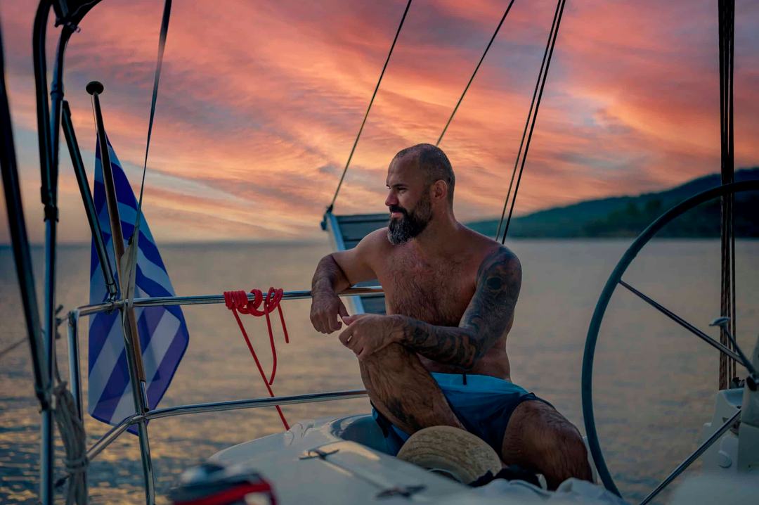 A shirtless bearded man with tattooed arms in blue swim shorts sitting at the stern of a sailing yacht beside the helm wheel and a Greek flag, under dramatic pink and orange sunset clouds