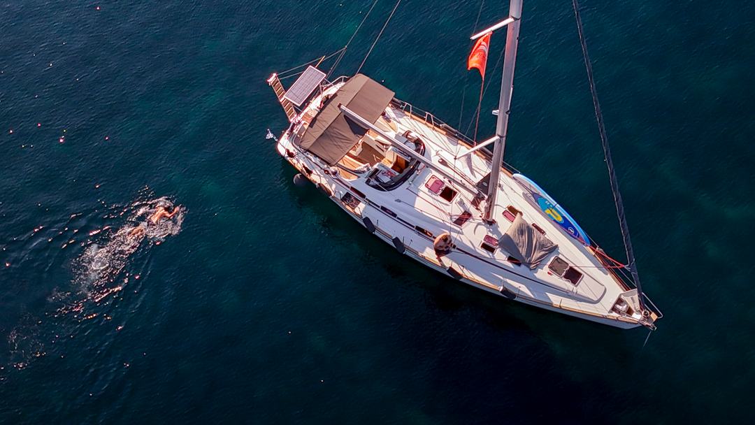 Aerial of a white sailing yacht with a tan bimini at anchor on dark blue-green water, a swimmer doing front crawl alongside