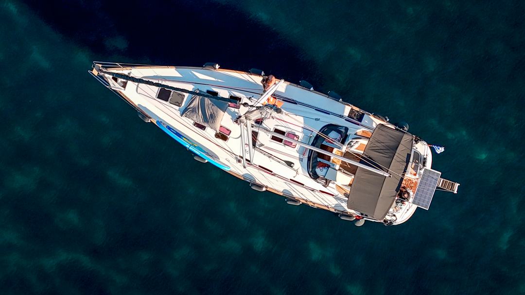 Top-down aerial of a white sailing yacht on dark teal water, its bimini, solar panel and gangway visible at the stern