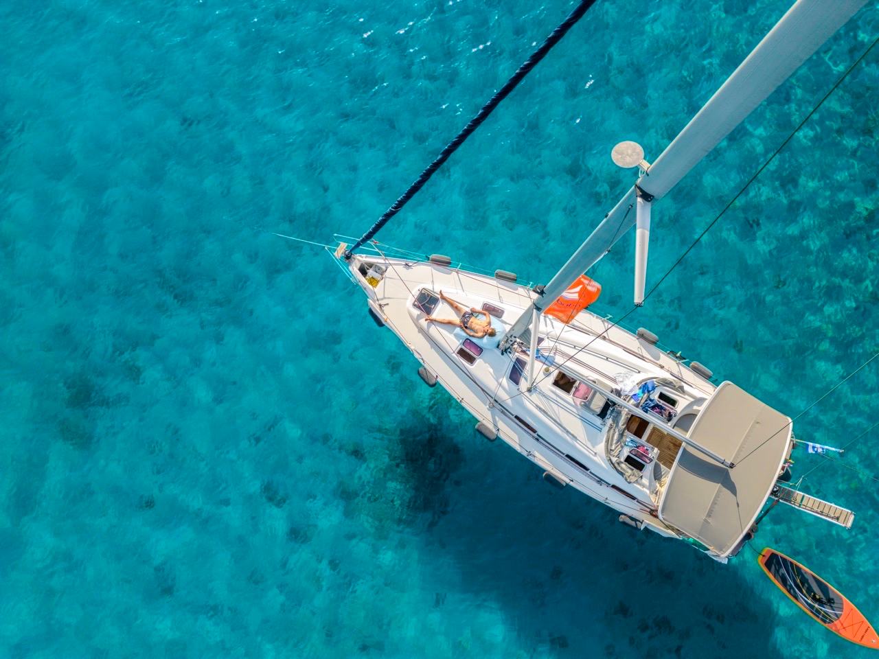 Top-down aerial of a sailing yacht over vivid turquoise shallows, a guest sunbathing on the foredeck and an orange paddleboard off the stern