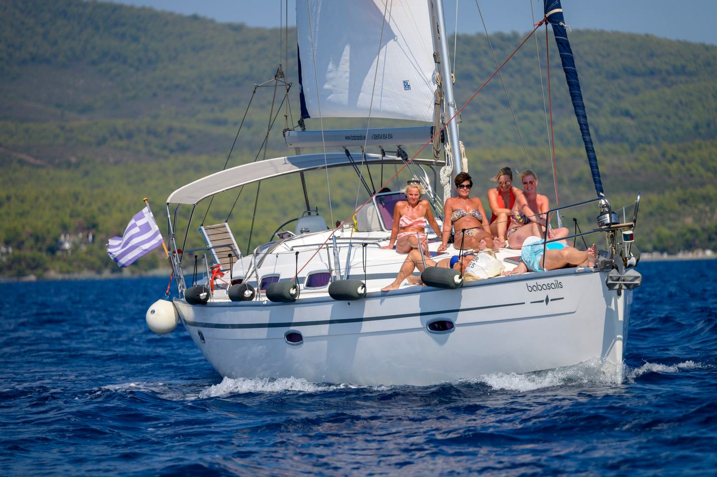 White Babasails sailing yacht under way with the mainsail partly raised, guests seated on the foredeck and pine-covered hills behind
